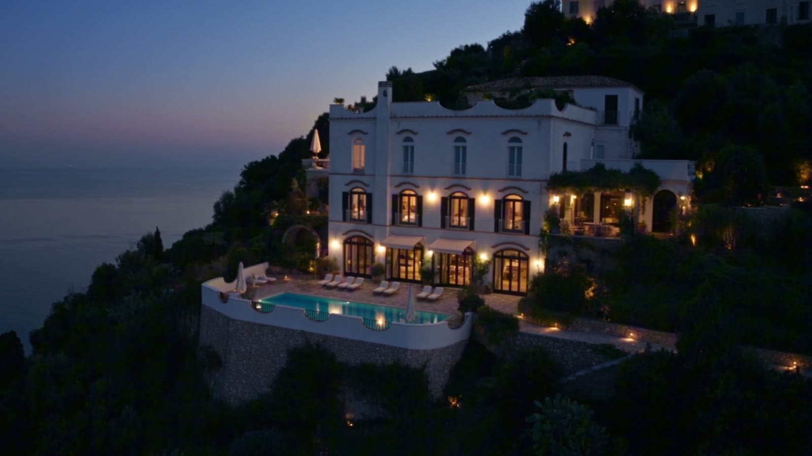 A clifftop villa with a lit infinity pool above the sea at dusk